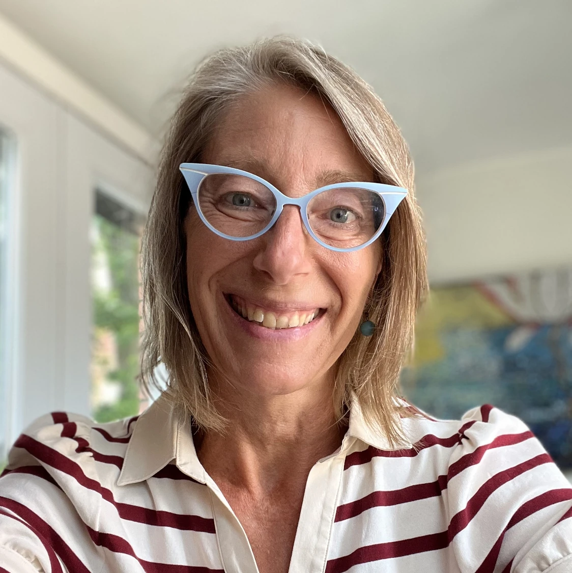A portrait of Jen wearing a striped shirt and smiling, standing in front of her office